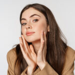 Beauty and skin care. Close up portrait of beautiful woman looking in mirror and touching her clear glowing face, standing over white background.