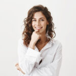 Emotive charming caucasian woman with curly hair and feather earring holding hand on ching and smiling broadly, expressing interest and positive attitude, standing against gray background. Copy space
